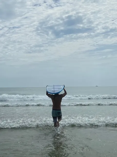 Student carrying a surfboard overhead, walking out into the water at Butler Beach